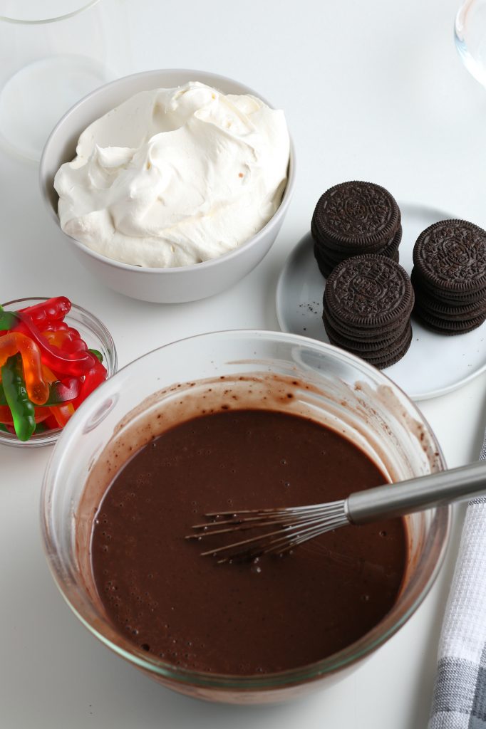 A bowl of chocolate pudding is surrounded by other dirt cups ingredients. 