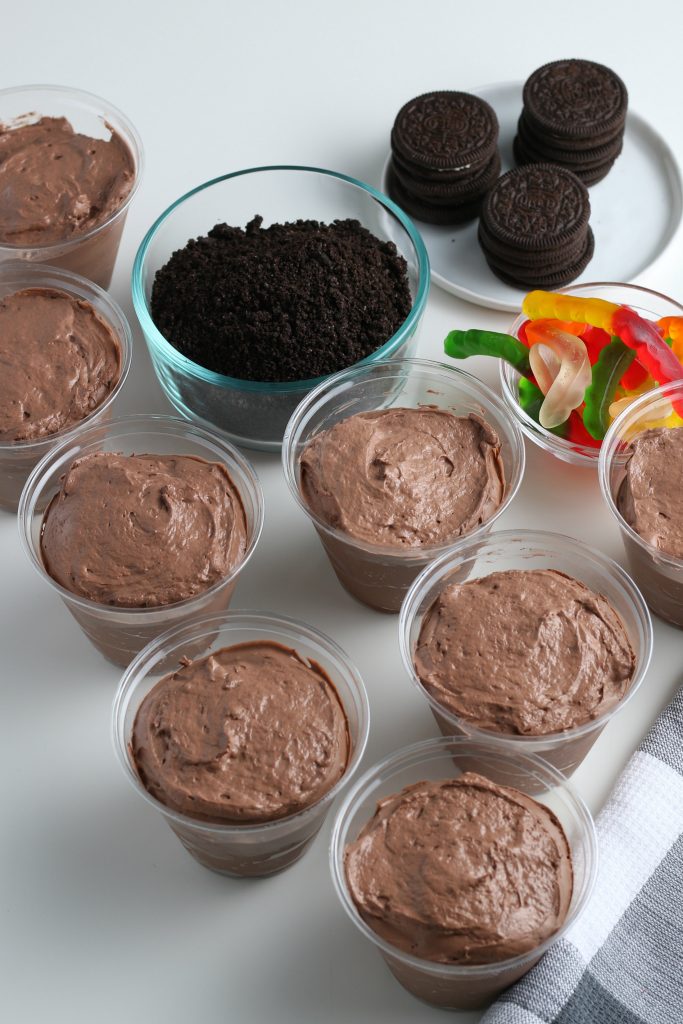 Little plastic cups of chocolate pudding are shown with a bowl of crushed Oreo cookies and another of gummy worms.