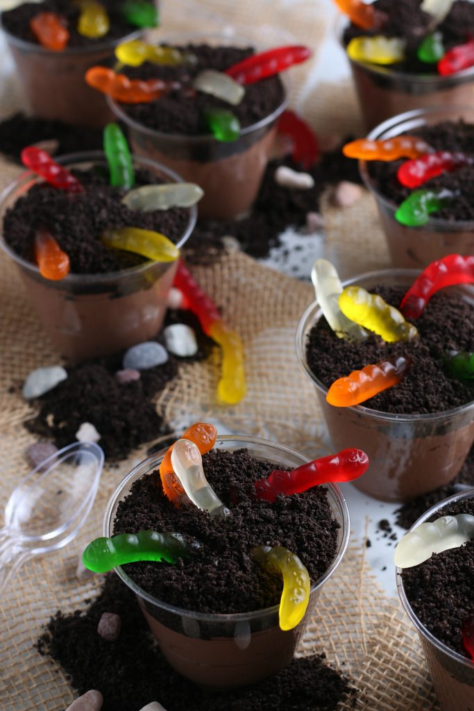 Burlap fabric is topped with plastic cups filled with chocolate pudding, crushed Oreo cookies and gummy worms. Plastic spoons sit nearby.