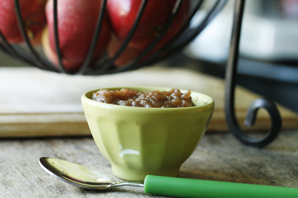 Homemade Honey Cinnamon Applesauce is shown in a green bowl on a wooden table with a spoon and a basket of apples nearby.