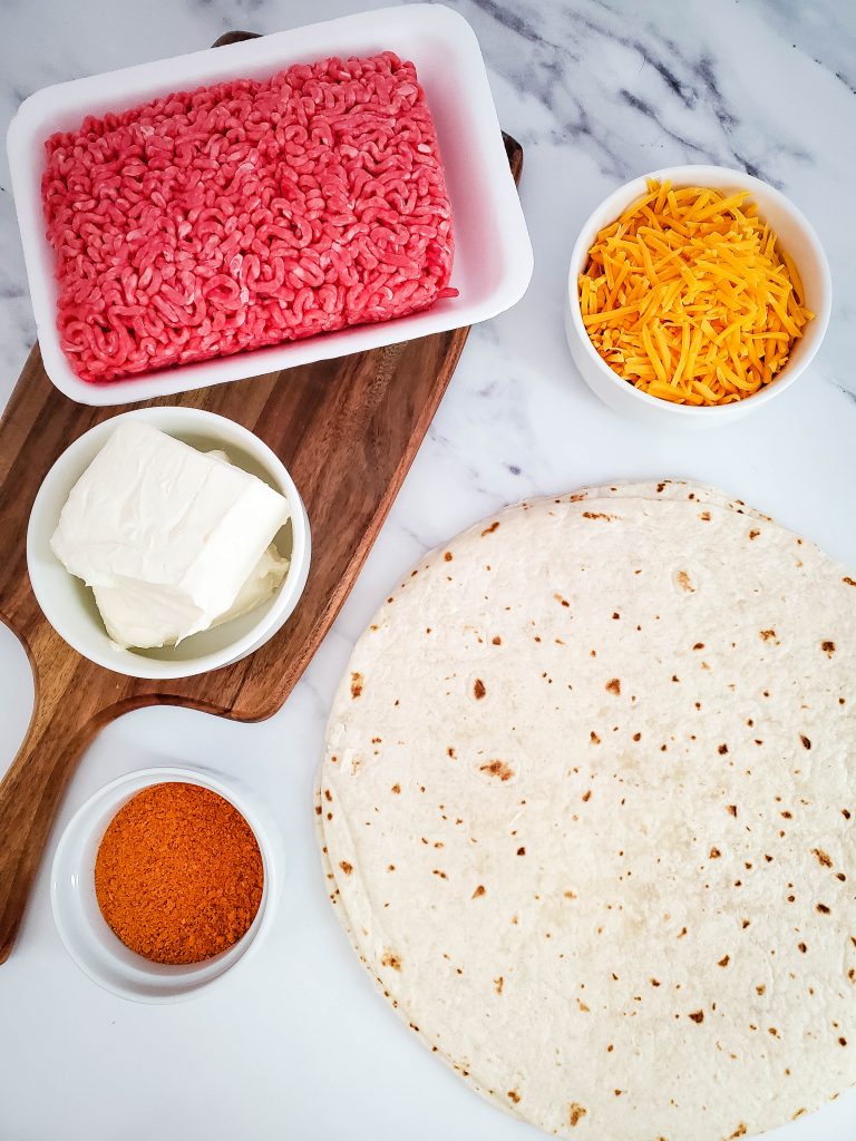 Ingredients including ground beef, shredded cheddar cheese, cream cheese and tortillas sit on a countertop.