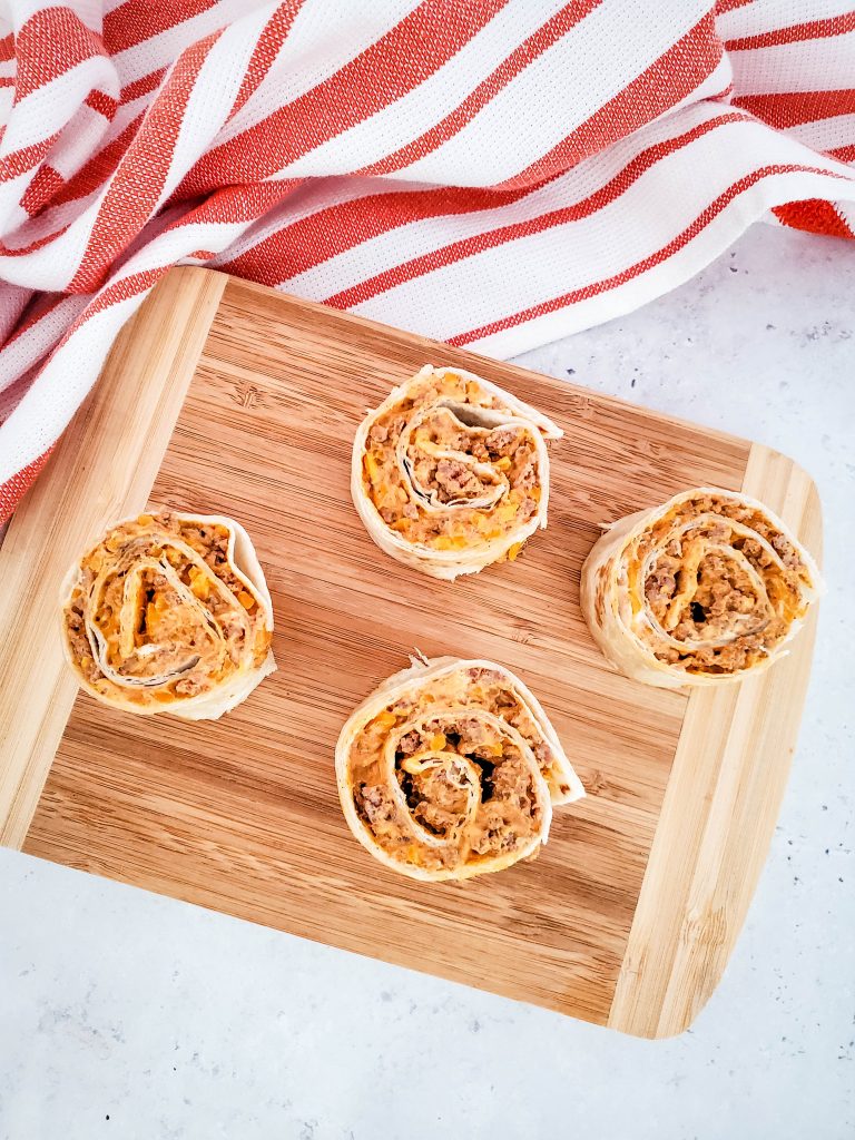 On a wooden cutting board, pinwheels filled with a cheesy beef filling sit. A red and white tea towel is arranged nearby.