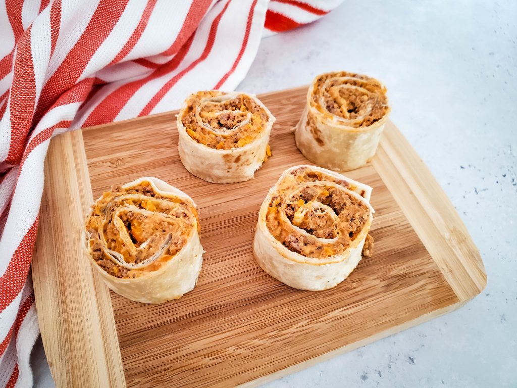 Cheesy Taco Pinwheels sit on a wooden cutting board with a red and white tea towel nearby.