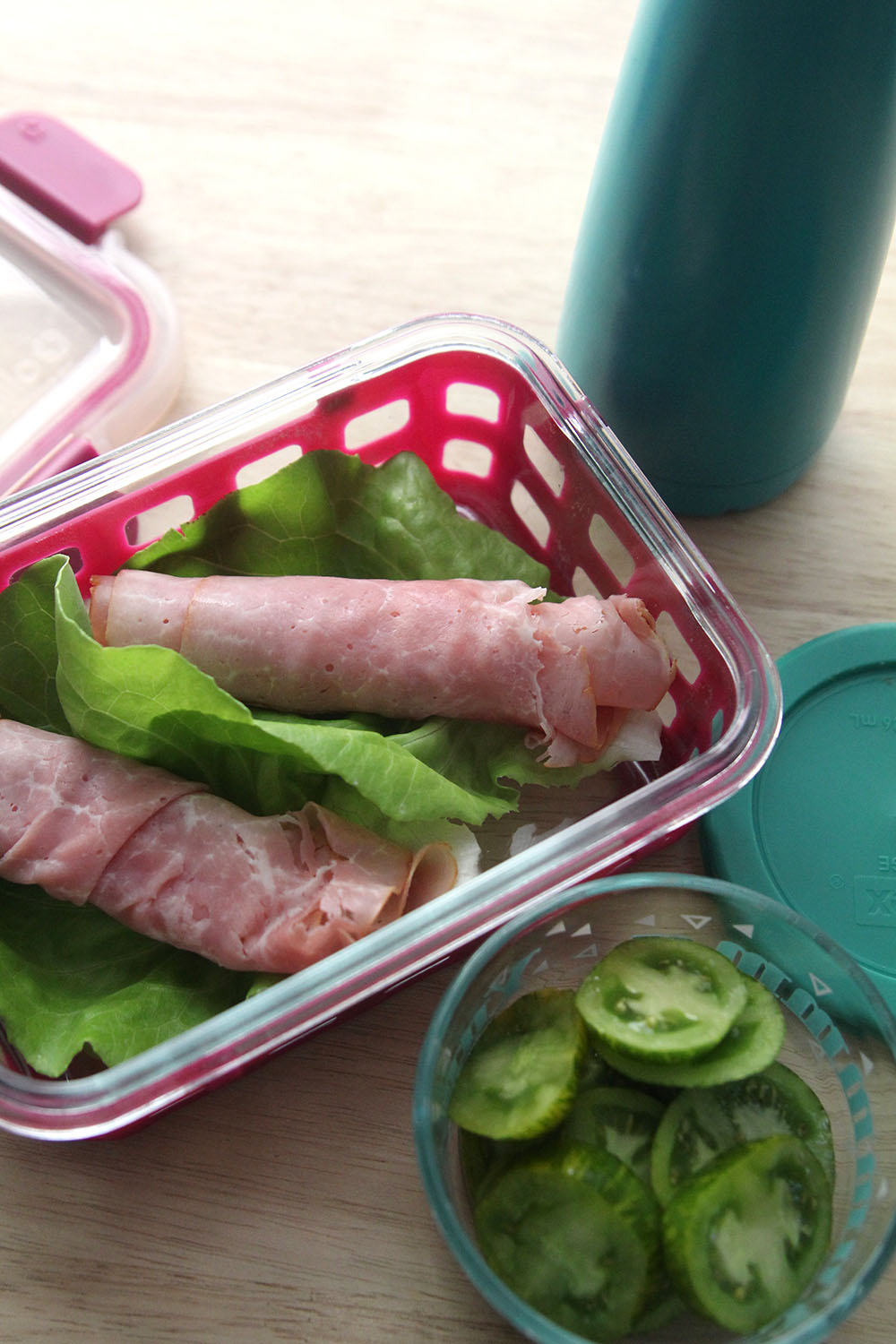 A glass container with two lettuce leaves topped with ham rolls is shown next to a glass container of green tomatoes, a water bottle and the container lids.