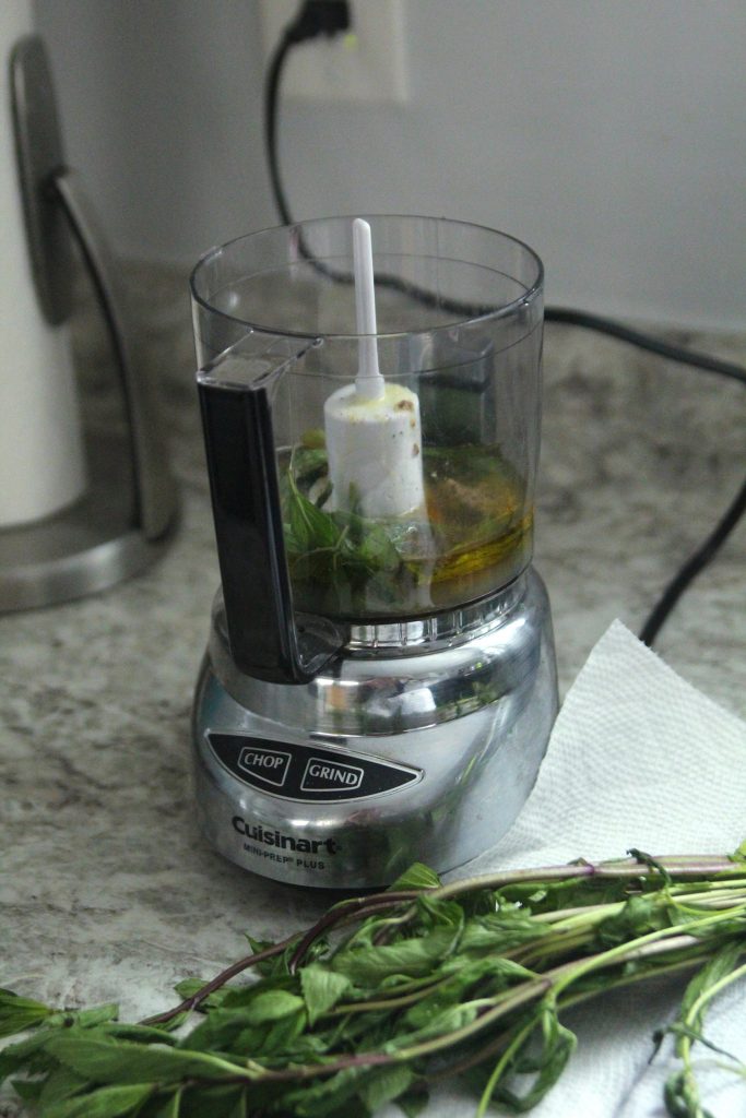 A small food processor is shown with mint leaves, oil and other ingredients inside. Nearby, fresh mint on stems sits on a white paper towel on the countertop.